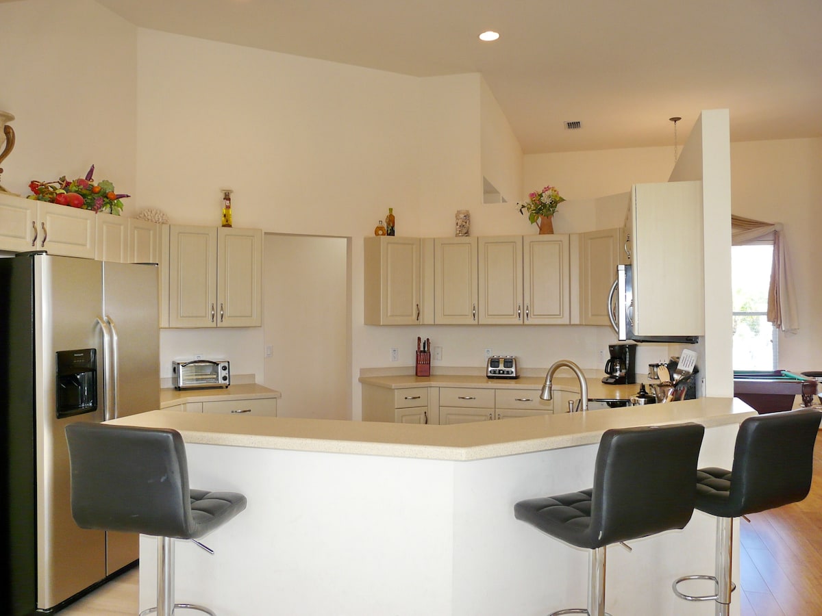 A modern kitchen with white cabinets, a stainless steel refrigerator, a built-in oven, a sink, and a bar area with two stools.