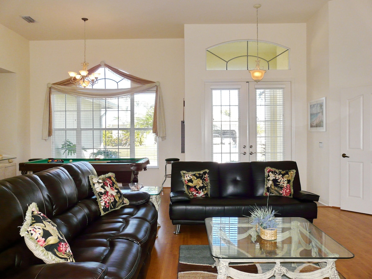 A spacious living room with a pool table, two leather sofas with floral cushions, a glass coffee table, and a chandelier.