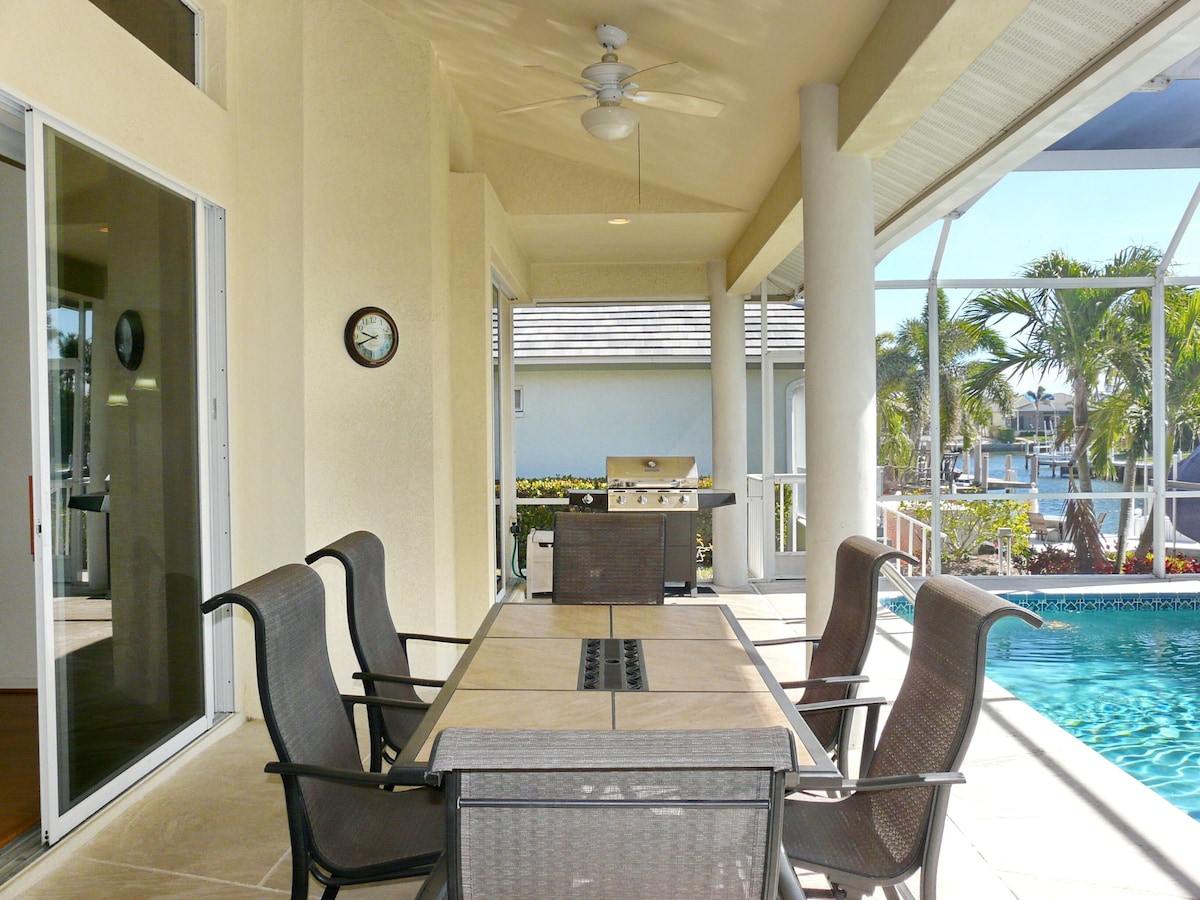 A covered outdoor dining area with a pool view, a grill, and a clock on the wall.