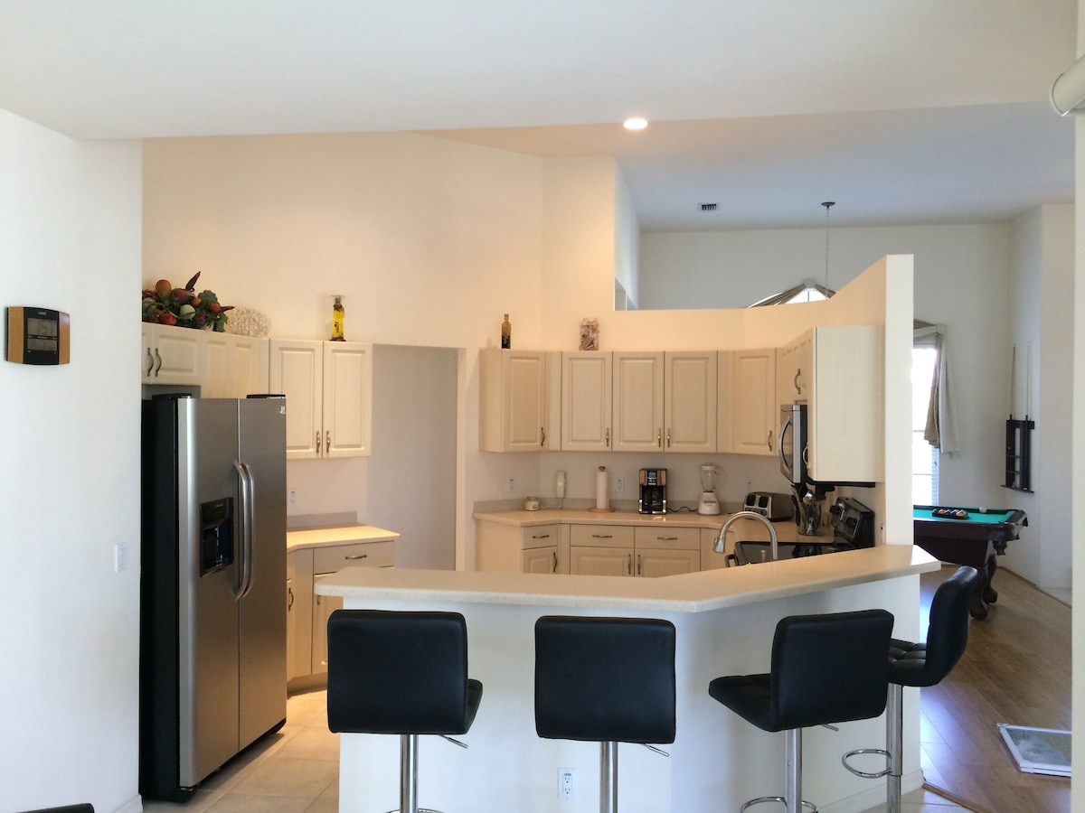 A modern kitchen with white cabinets, a stainless steel refrigerator, a built-in island with bar stools, and a pool table in the adjacent room.