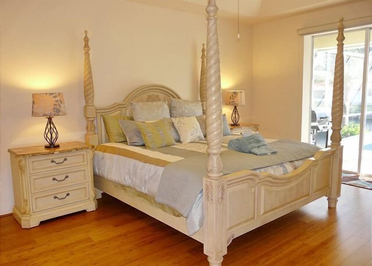 A four-poster bed with a wooden headboard, a bedside table with a lamp, and a window with a view of the outdoors.