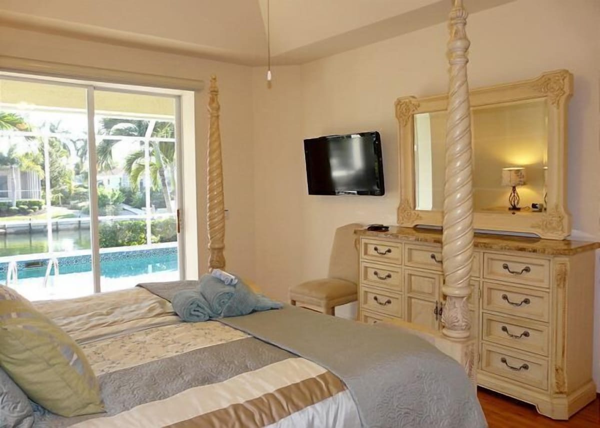 A bedroom with a four-poster bed, a television, a dresser, and a view of a pool.