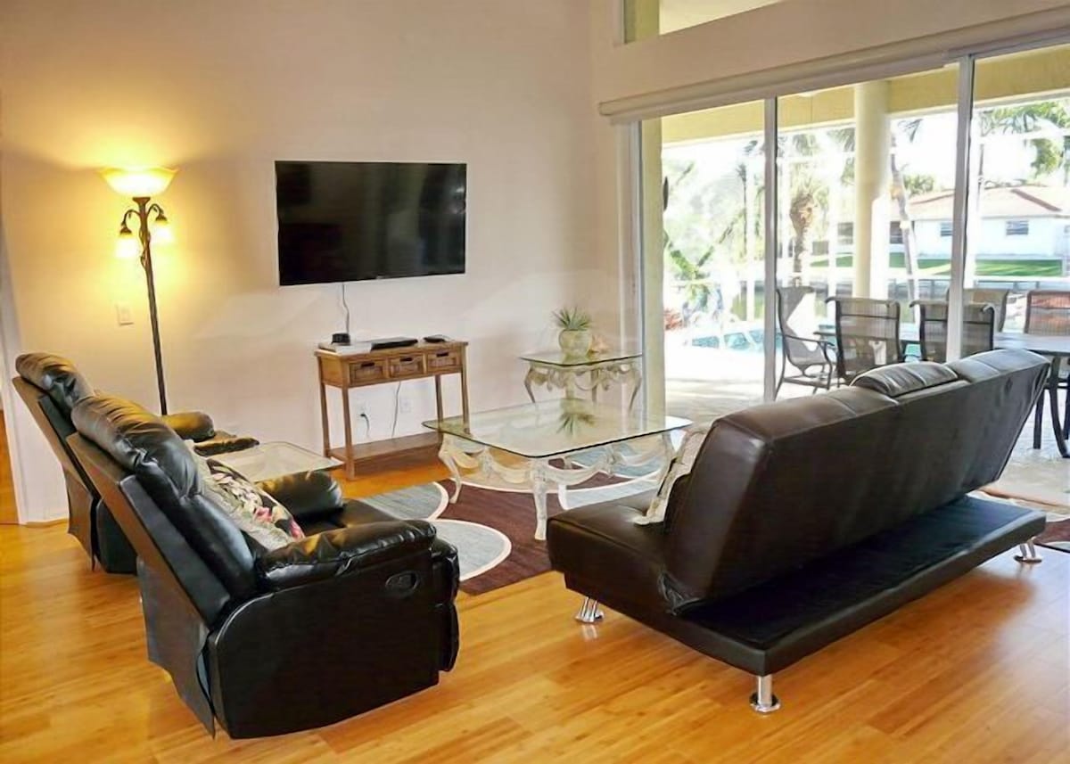A living room with a sofa, a coffee table, and a TV. There is a glass table with a plant on it, and a wooden side table. The room has large windows with a view of the outside.