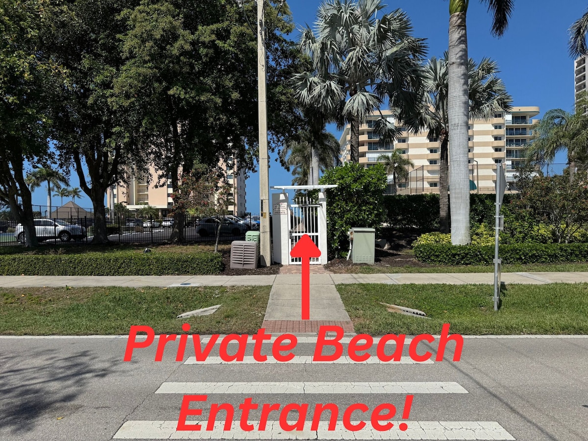 A private beach entrance with a clear pathway leading to a gated area, surrounded by palm trees and residential buildings.