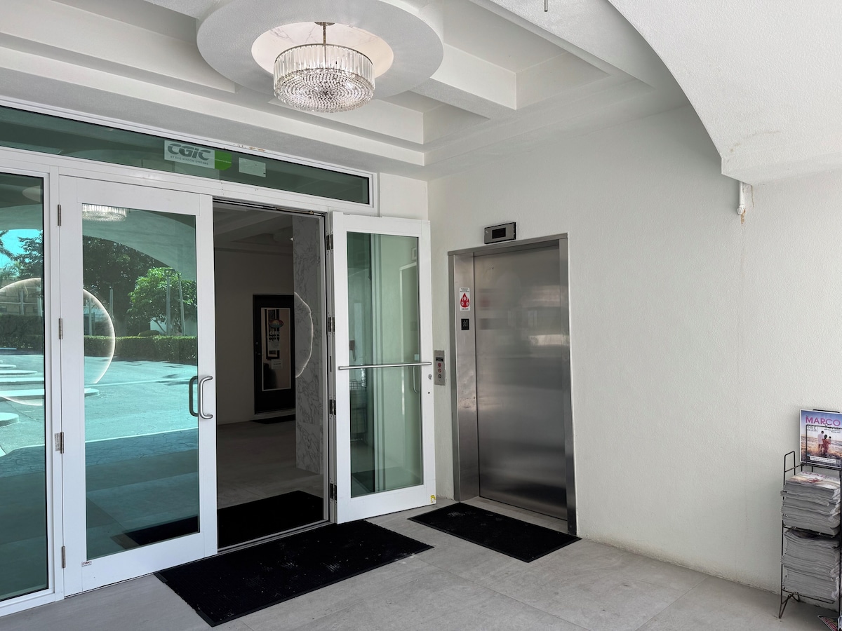 A hotel entrance with a glass door, a reception desk, a digital elevator sign, and a magazine rack.