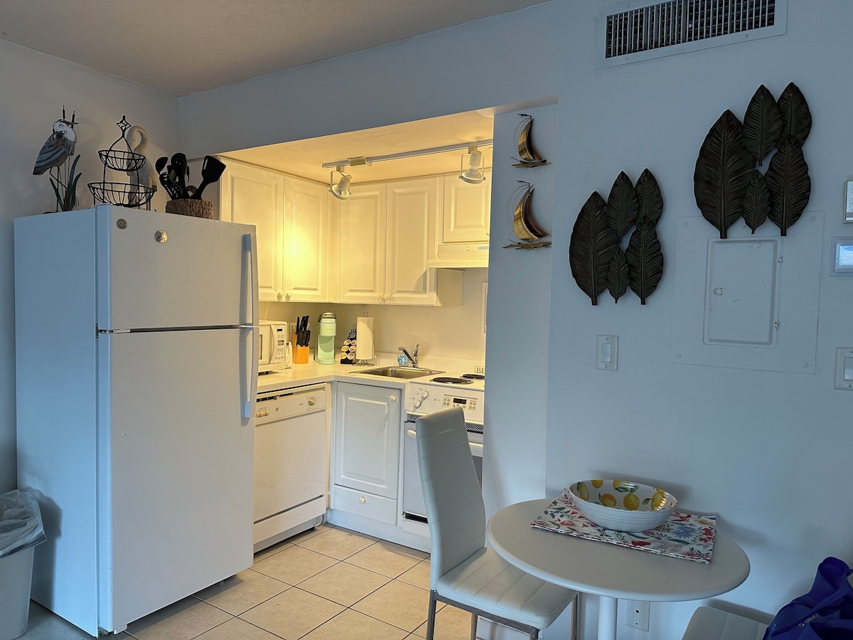 A compact kitchen with white cabinets, a refrigerator, a dining table with chairs, and decorative wall art.