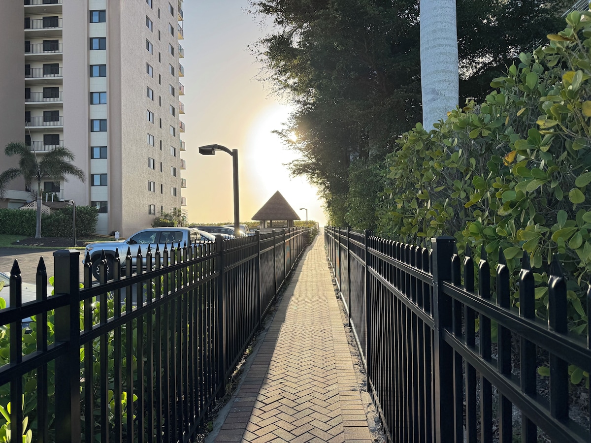 A paved walkway flanked by a black metal fence, leading to a building with a triangular roof structure.