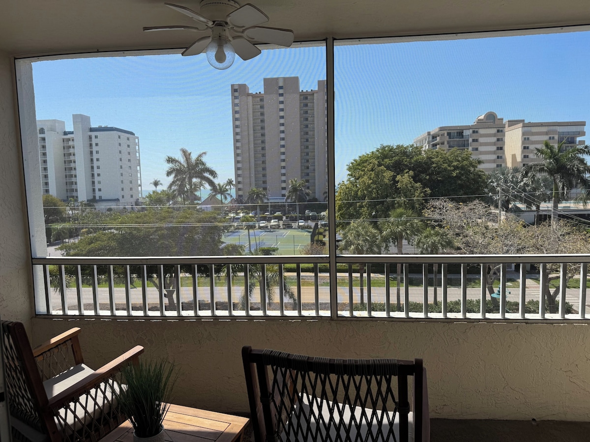 A balcony with a view of tall buildings, a tennis court, and a palm tree.