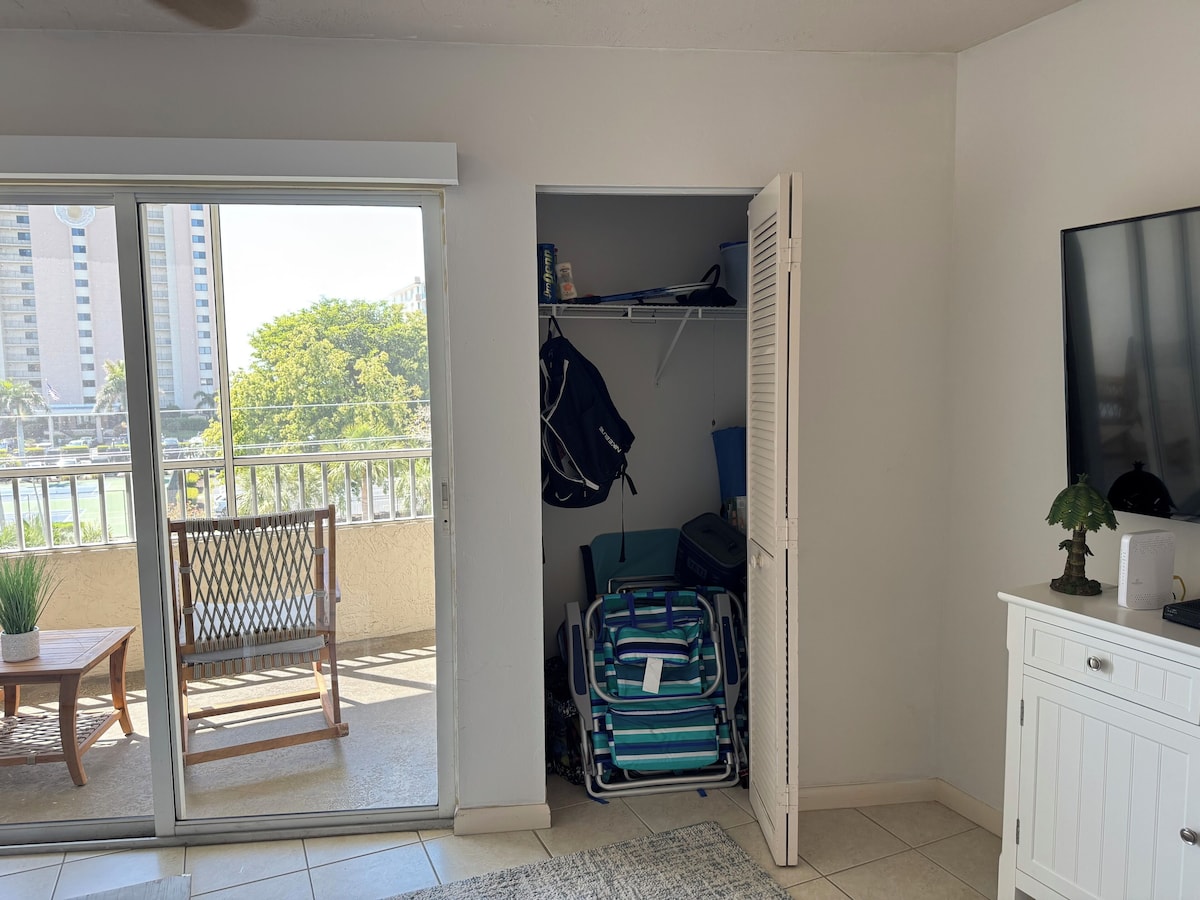 A room with a sliding glass door to a balcony, a small table with a plant, a wicker chair, a closet with hanging clothes and a suitcase, and a TV on a cabinet.