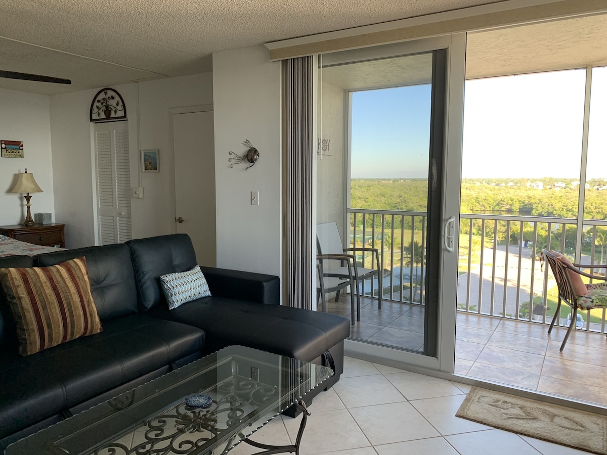 A living room with a black leather sofa, a glass coffee table, and a sliding glass door leading to a balcony with a view of greenery.