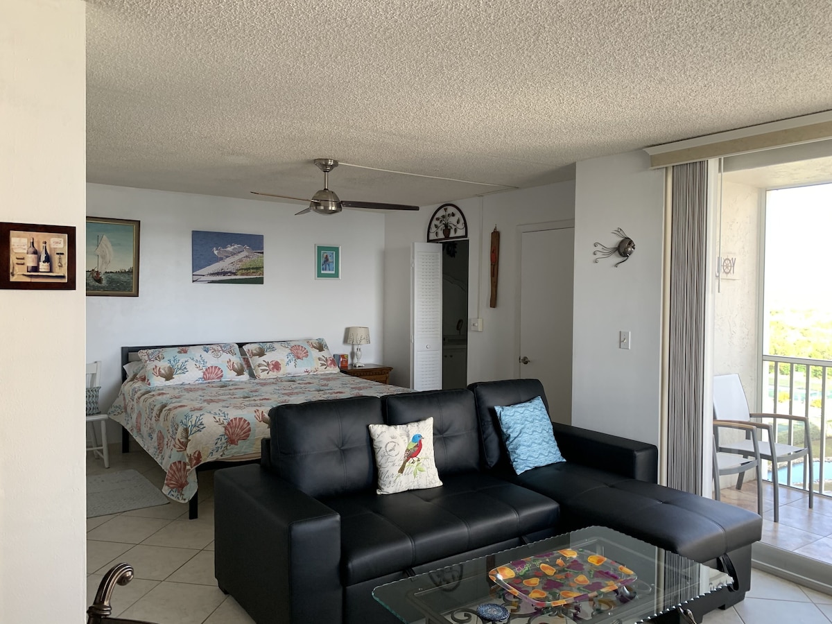 A living room with a black leather sofa, a glass coffee table, and a bed with floral bedding.