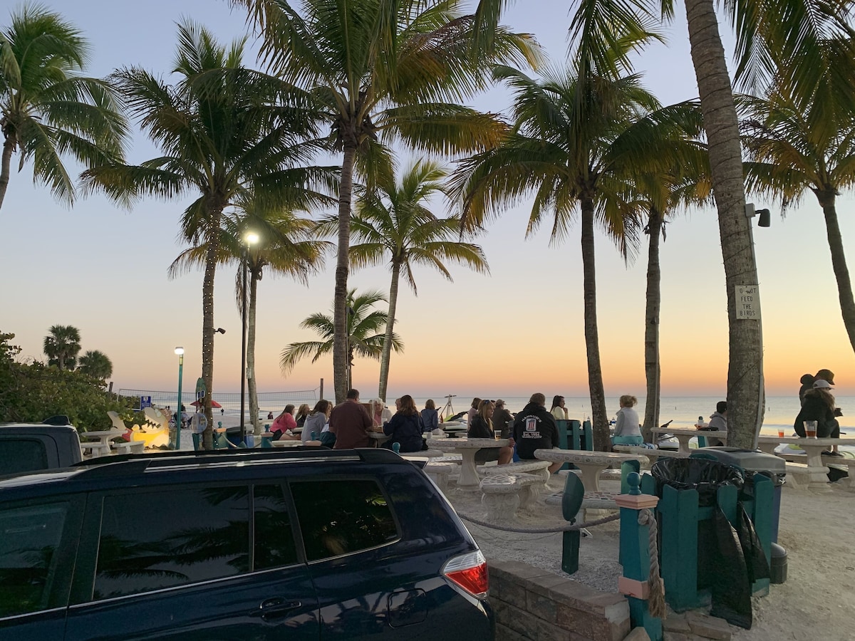 A beachside area with palm trees, picnic tables, and a car parked nearby.