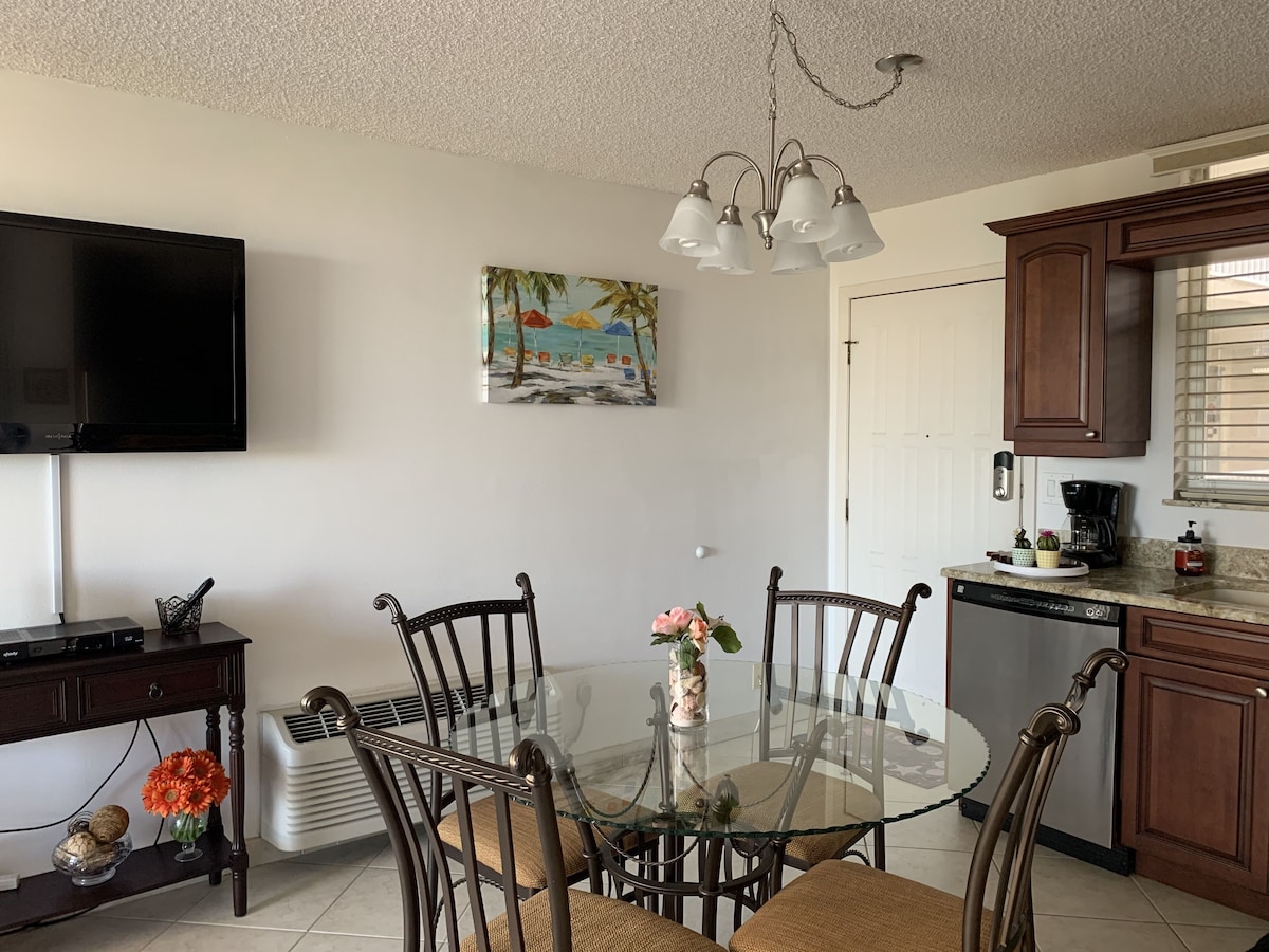 A dining area with a glass table surrounded by chairs, a television mounted on the wall, and a painting of a beach scene.