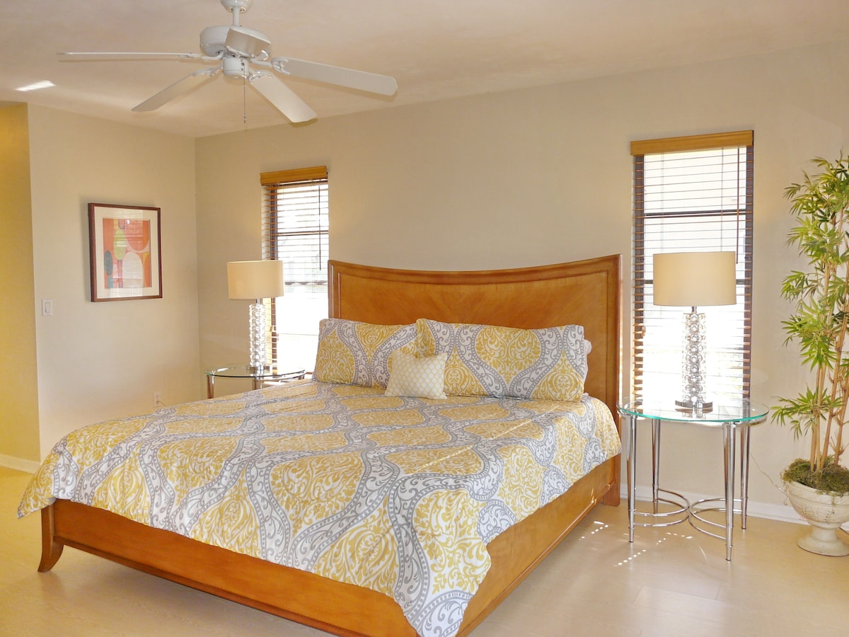 A bedroom with a wooden bed, two bedside lamps, a ceiling fan, and a potted plant.