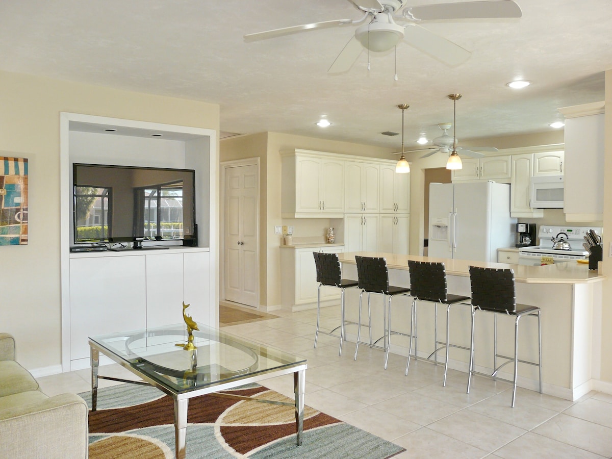 A modern kitchen with white cabinets, a central island with bar stools, a glass-top dining table, and a ceiling fan.