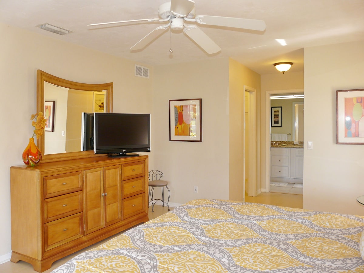 A bedroom with a bed, a wooden dresser, a television on the dresser, a ceiling fan, and a bathroom visible through an open door.