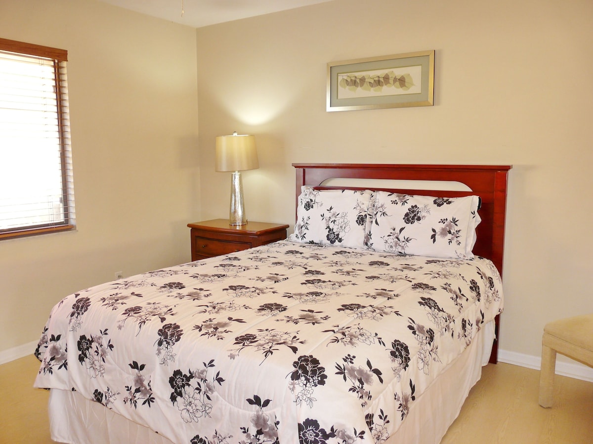 A bedroom with a bed, a nightstand, a lamp, a chair, and a framed picture on the wall.