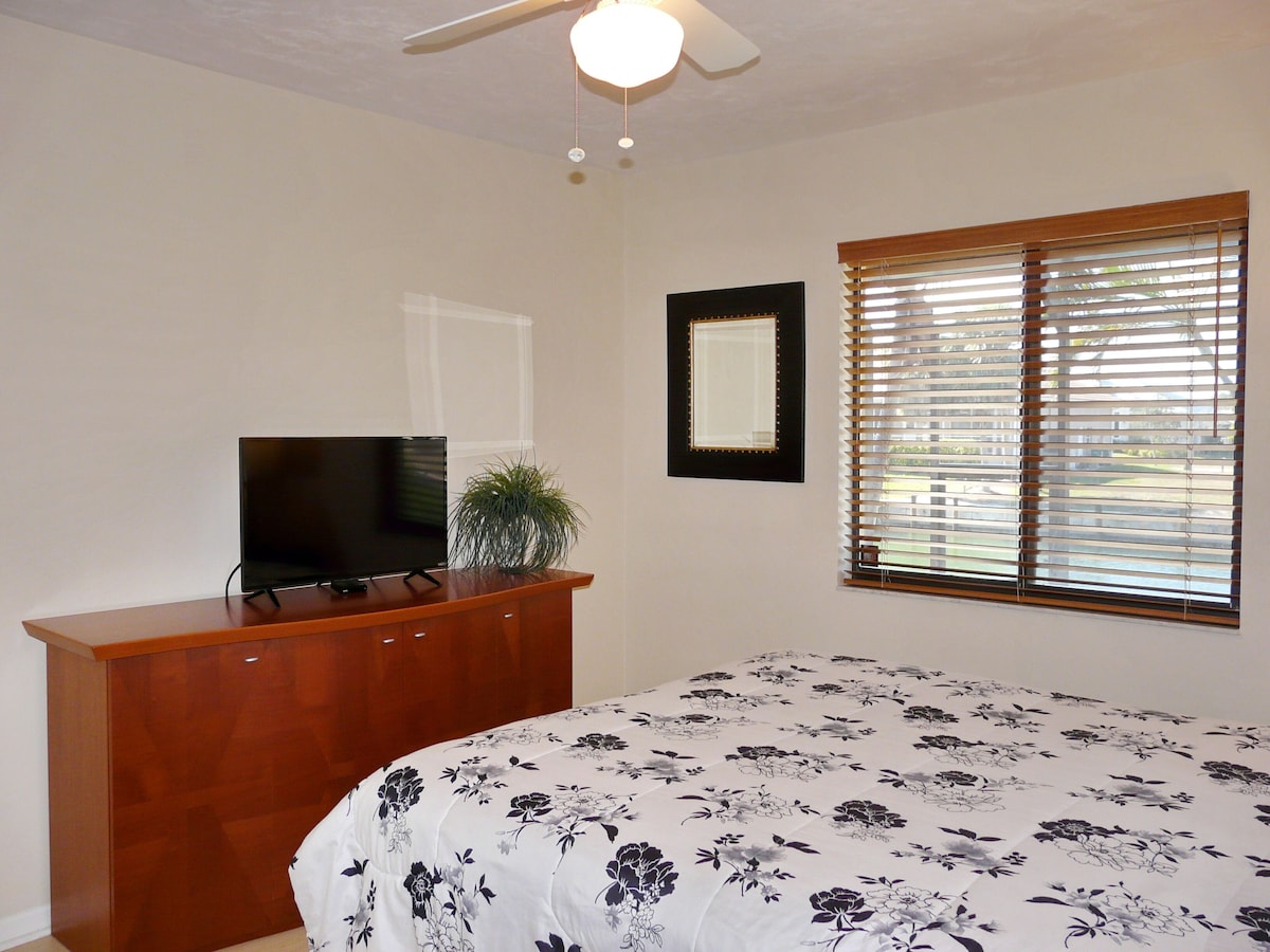 A bedroom with a bed, a TV on a wooden dresser, a framed picture on the wall, and a window with blinds.