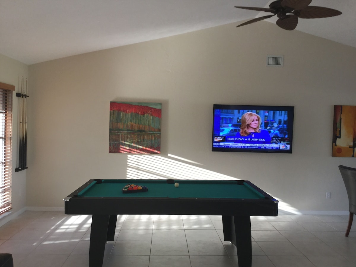 A pool table with balls, a television displaying a news program, and a painting on the wall.