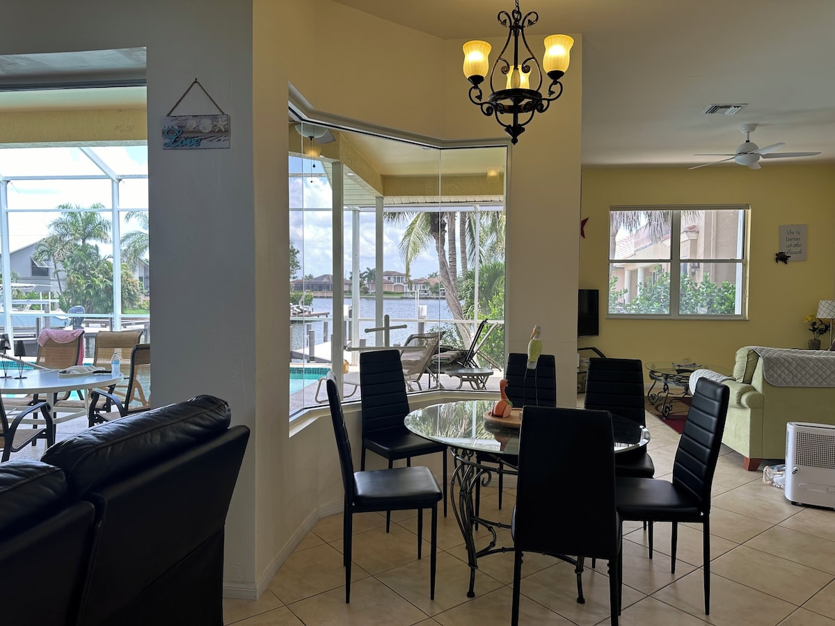 A dining area with a view of the pool and palm trees, a chandelier, and a ceiling fan.
