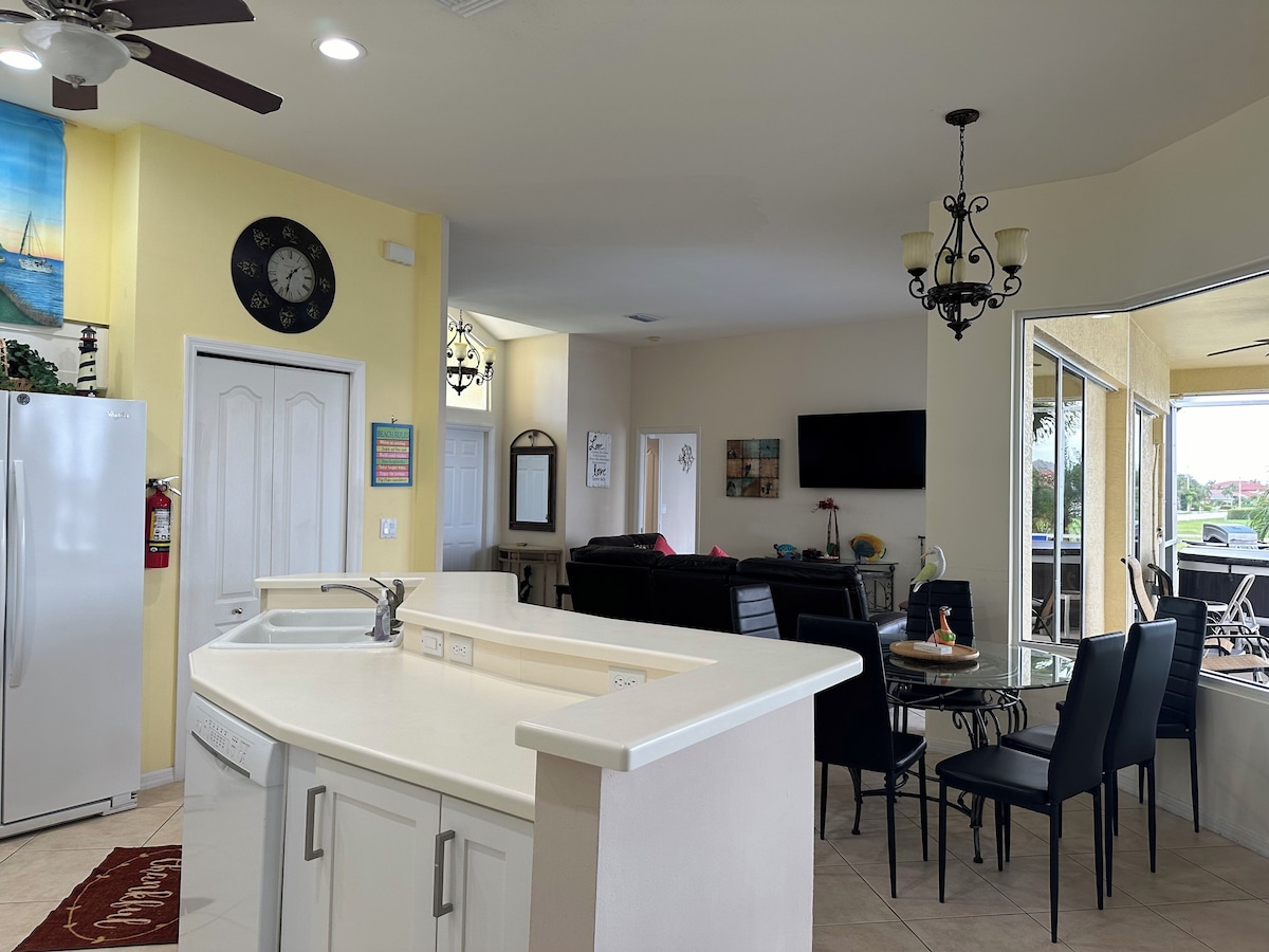 A kitchen with a white countertop, a dining area with black chairs, a ceiling fan, and a mounted TV.
