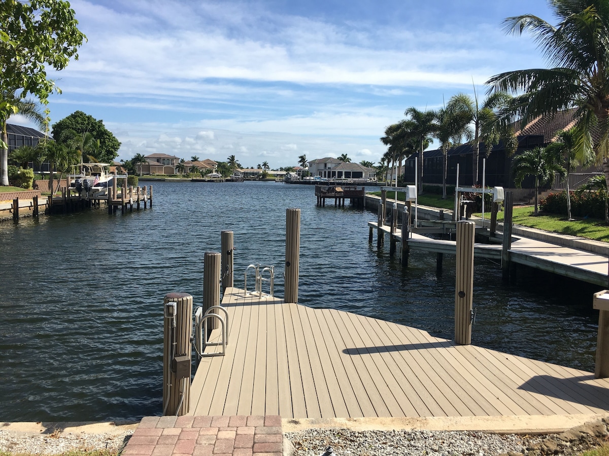 A wooden dock extending into a calm body of water, surrounded by residential buildings and palm trees.