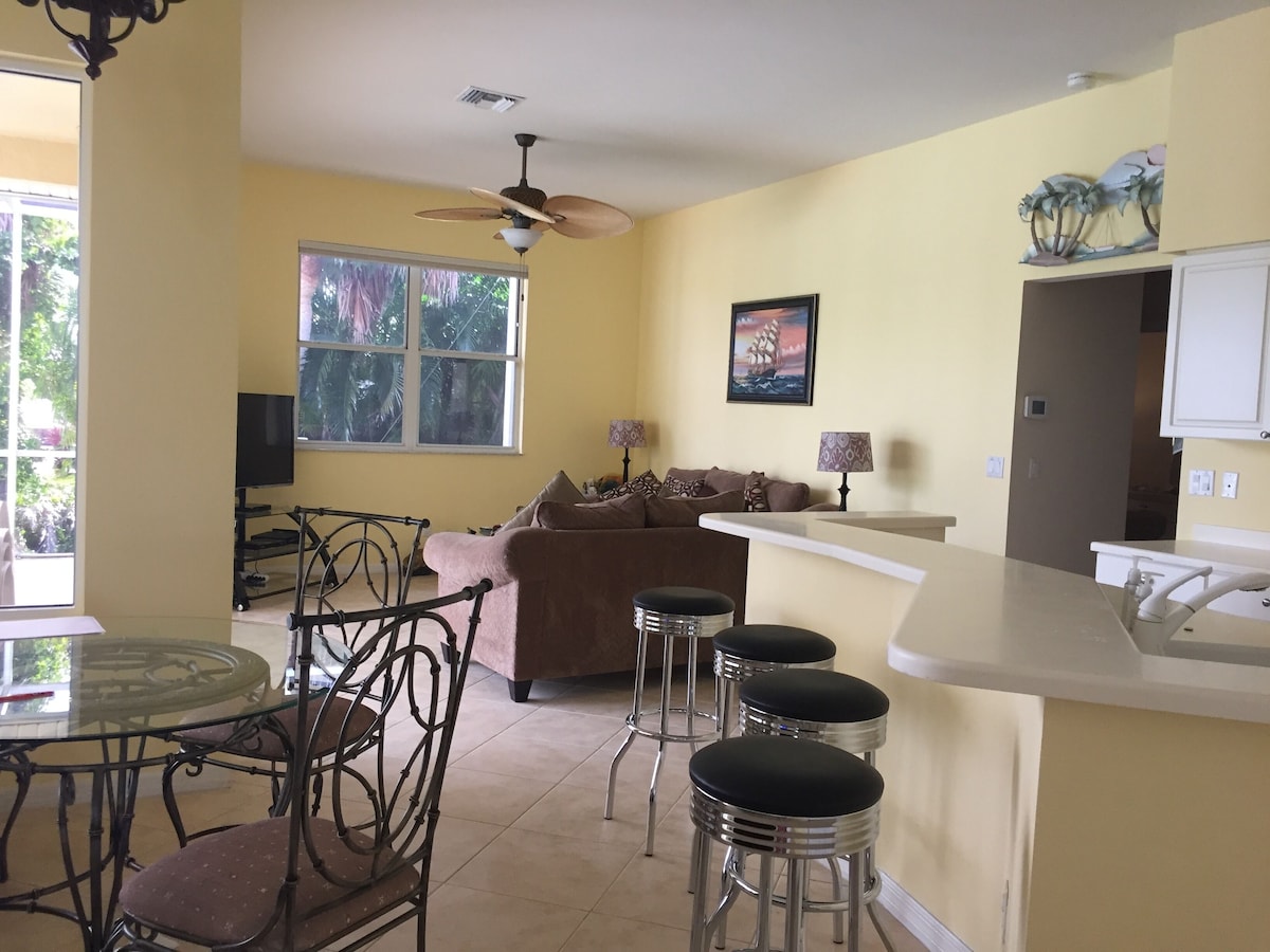 A compact kitchen with a dining area, a ceiling fan, a large window, and a sofa.