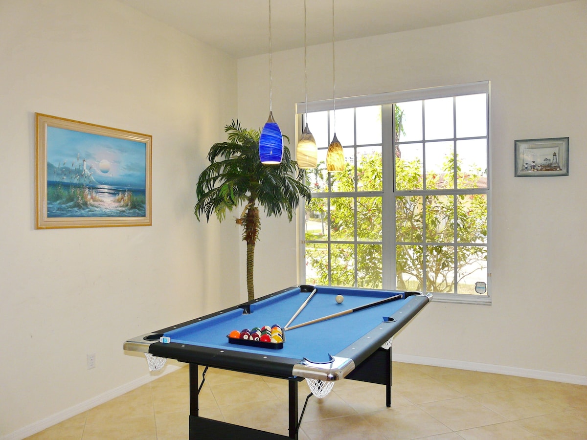 A pool table with balls, a framed painting, and a window with a view of greenery.