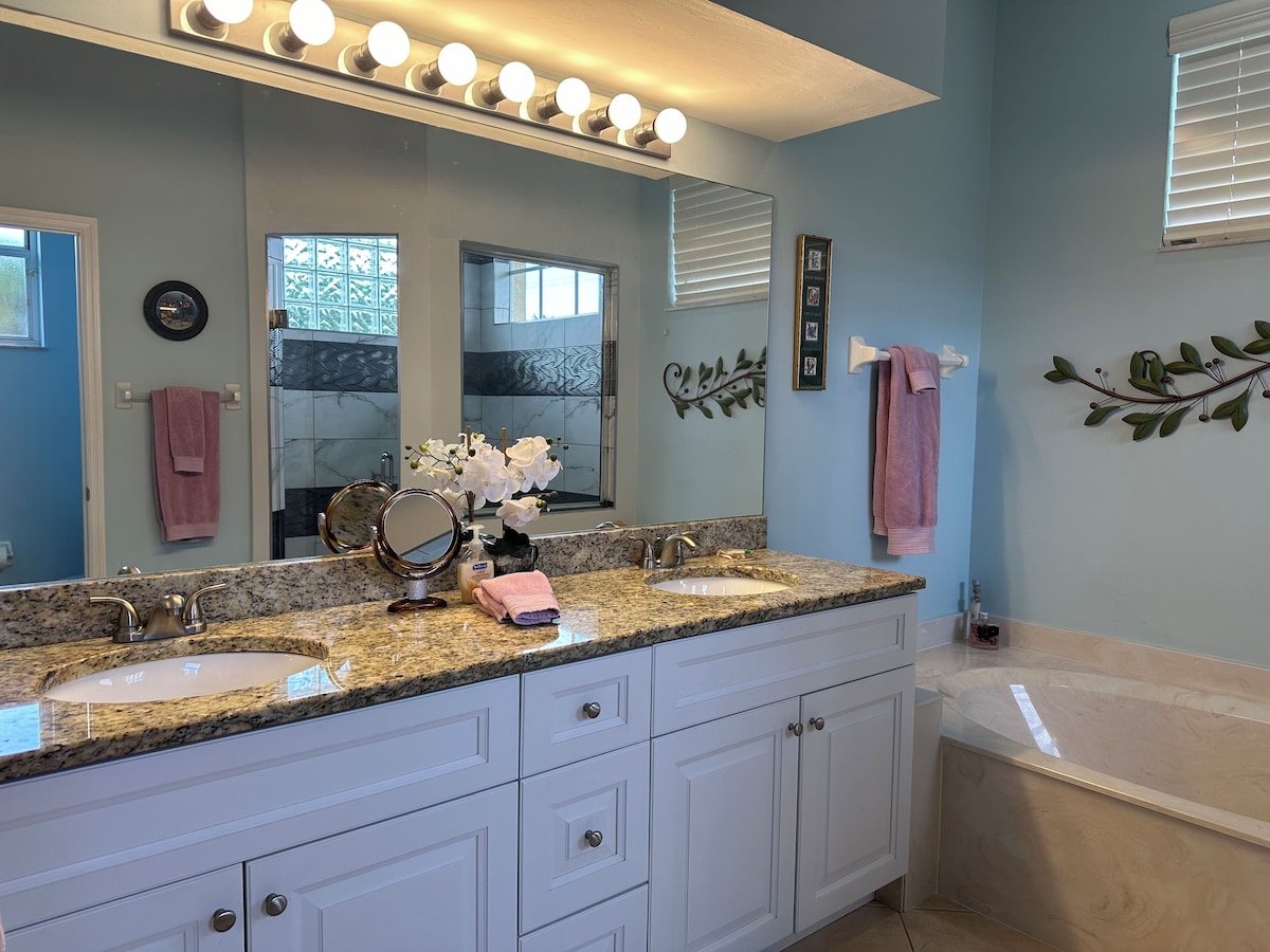A bathroom with a double sink vanity, a large mirror with lights, a towel rack, and a decorative plant.