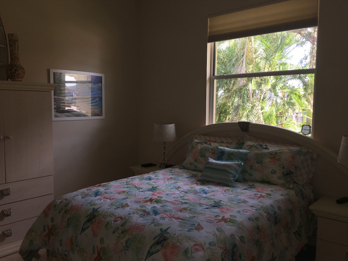A bedroom with a bed, a nightstand, a lamp, a dresser, a framed picture on the wall, and a window with a view of palm trees.