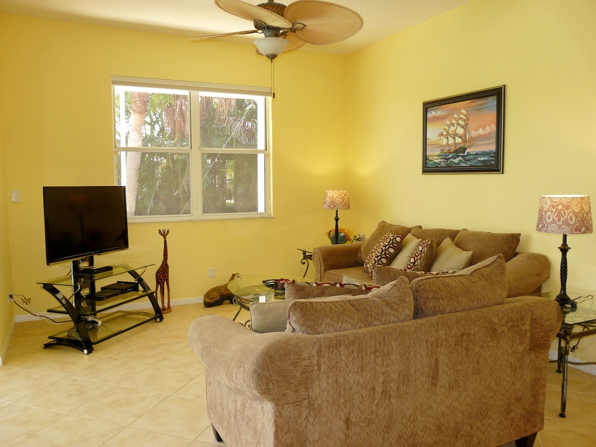 A living room with a beige sofa, a flat-screen TV, a ceiling fan, and a window with a view of trees.