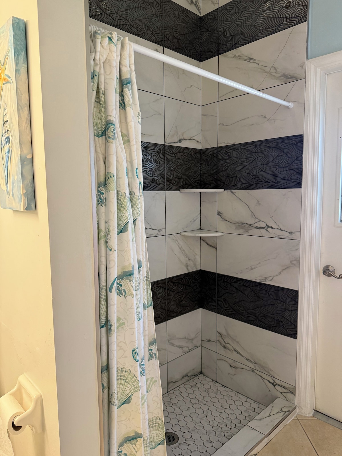 A shower area with a white curtain featuring a shell pattern, a white door, and a white towel rack.