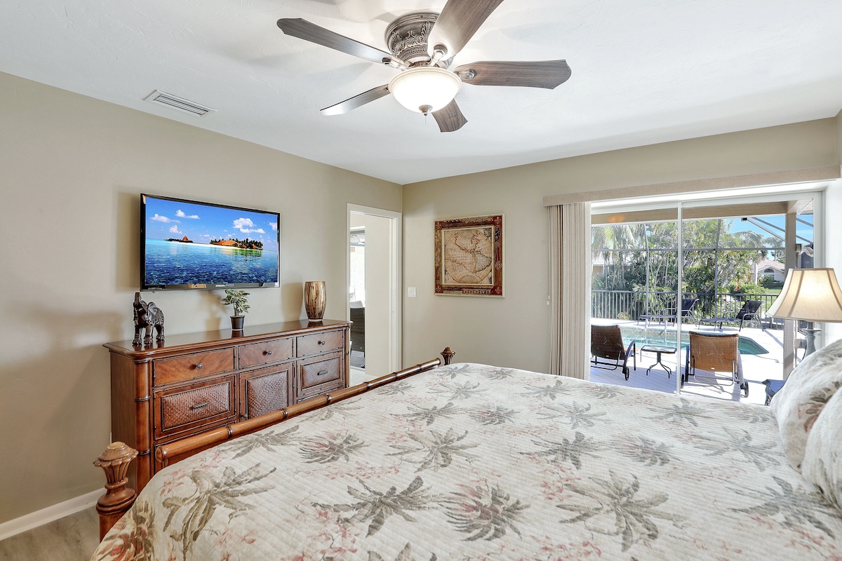 A bedroom with a bed, a ceiling fan, a television, a dresser, and a view of a pool and outdoor seating.