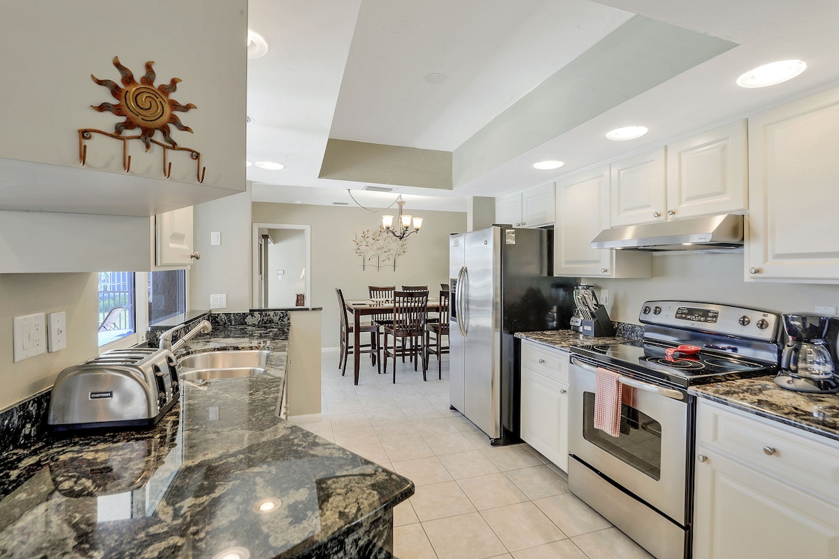A modern kitchen with granite countertops, stainless steel appliances, and a dining area with wooden chairs.