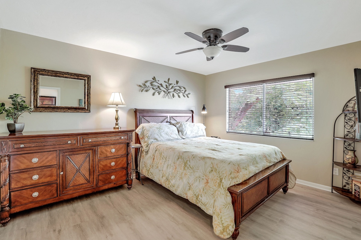 A bedroom with a wooden bed, a ceiling fan, a large window with blinds, a dresser, and a mirror.