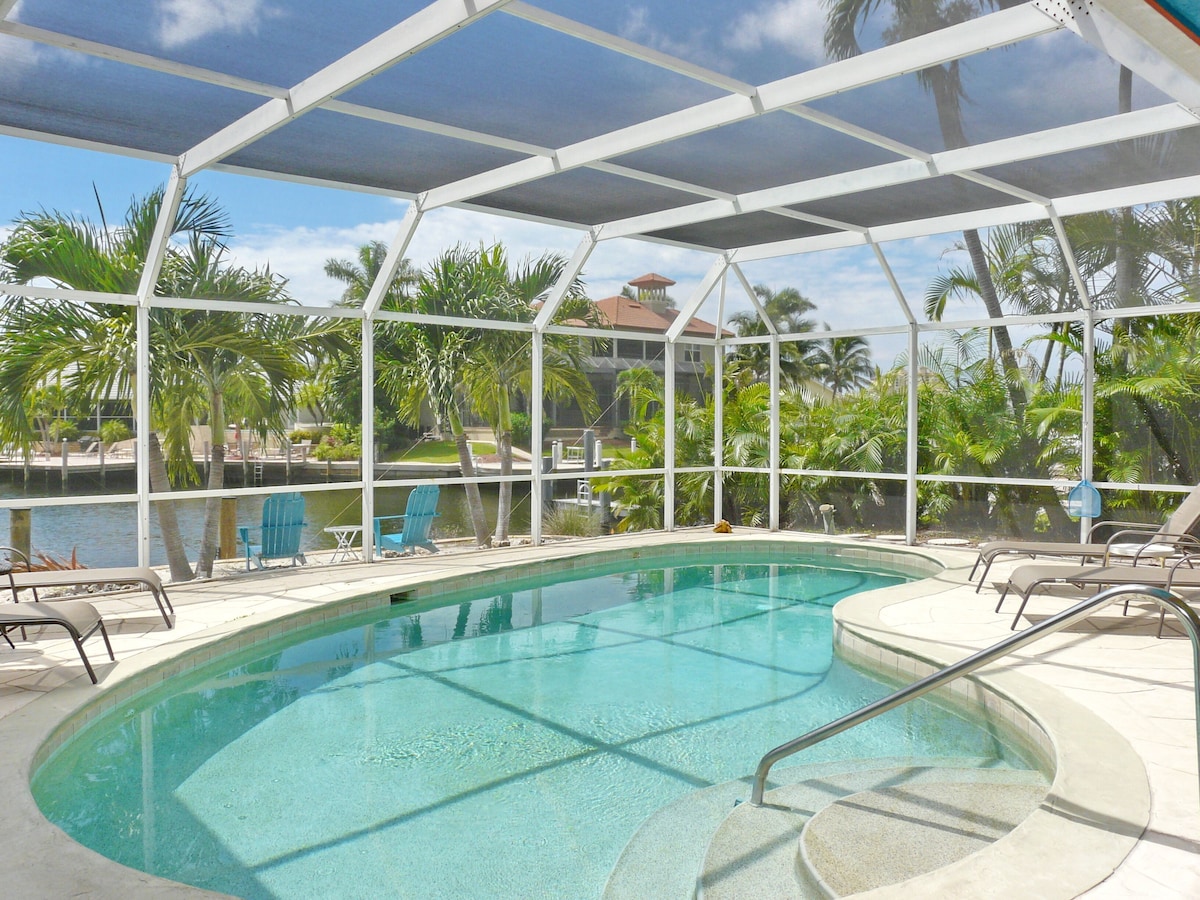An indoor swimming pool with clear blue water, surrounded by palm trees and a covered area with chairs.