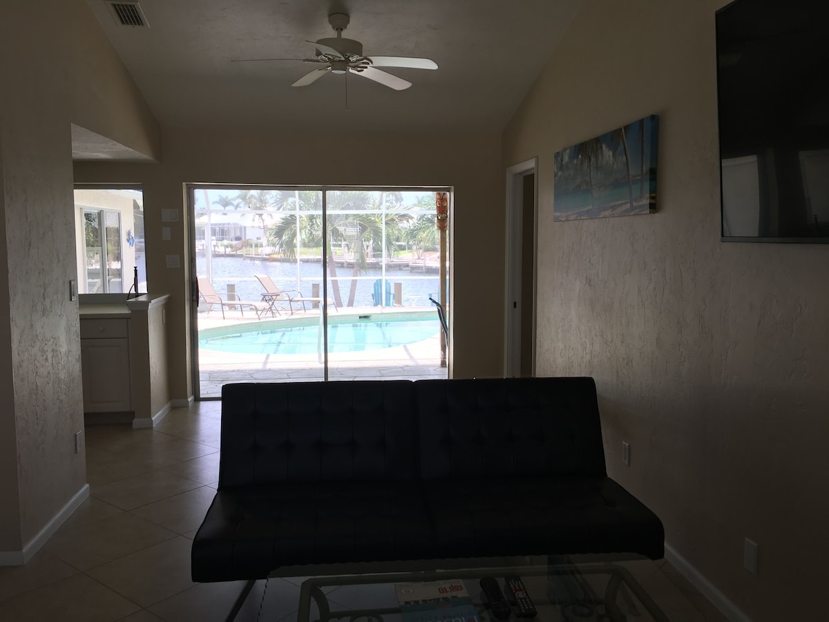A living room with a sofa, a ceiling fan, a glass table with remote controls, and a view of a pool through glass doors.
