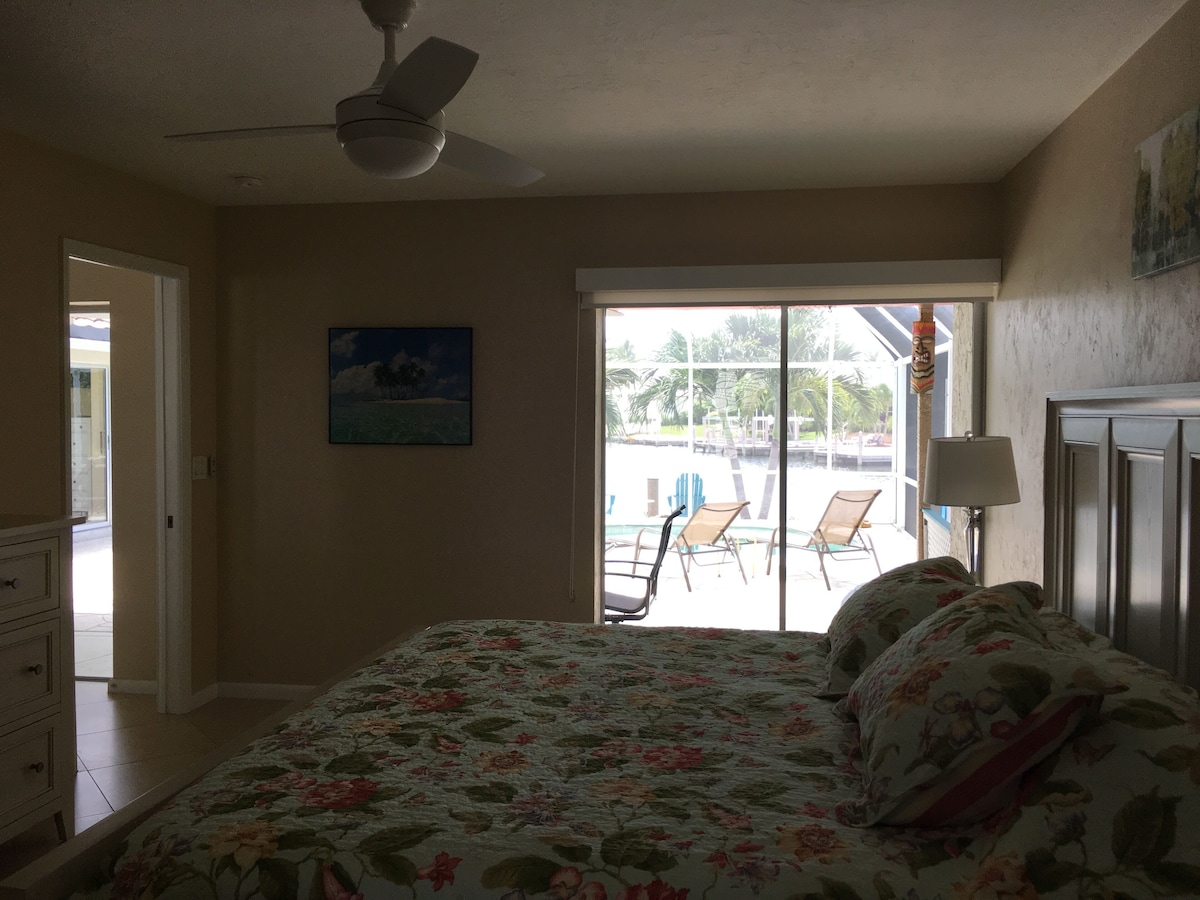 A bedroom with a bed, a ceiling fan, a painting, a view of a pool and chairs, and a door leading to another room.