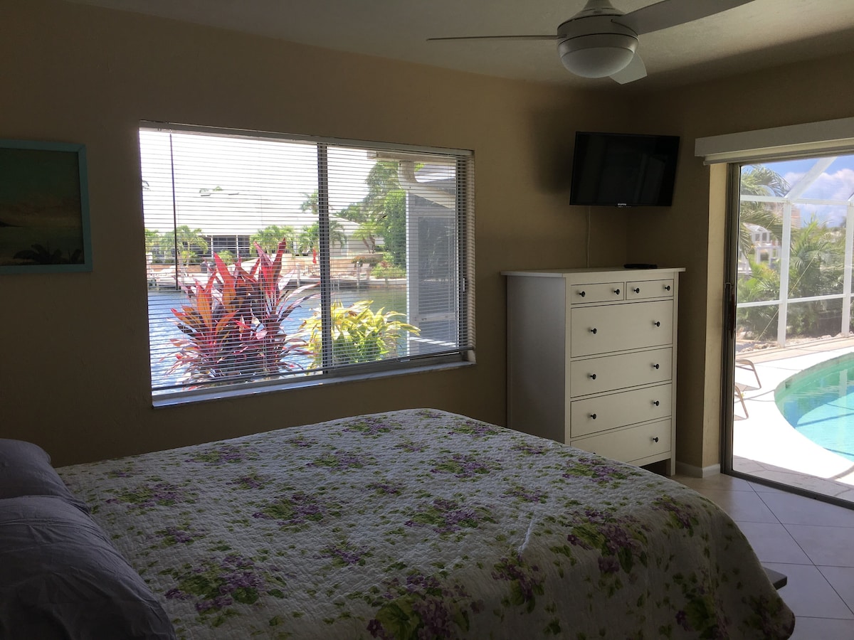 A bedroom with a bed, a dresser, a ceiling fan, a TV, and a window with a view of a swimming pool and palm trees.