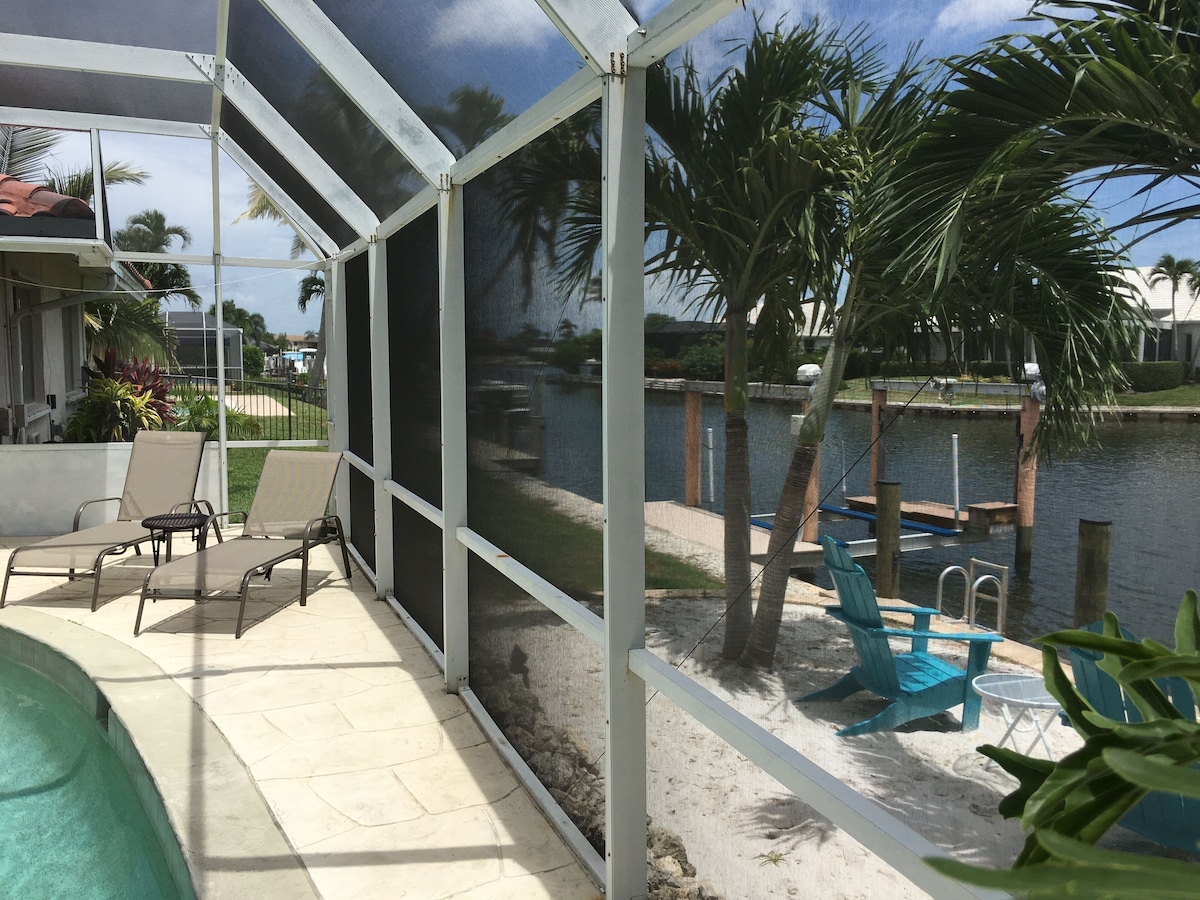 A covered outdoor area with lounge chairs, a pool, and a view of a body of water with palm trees.
